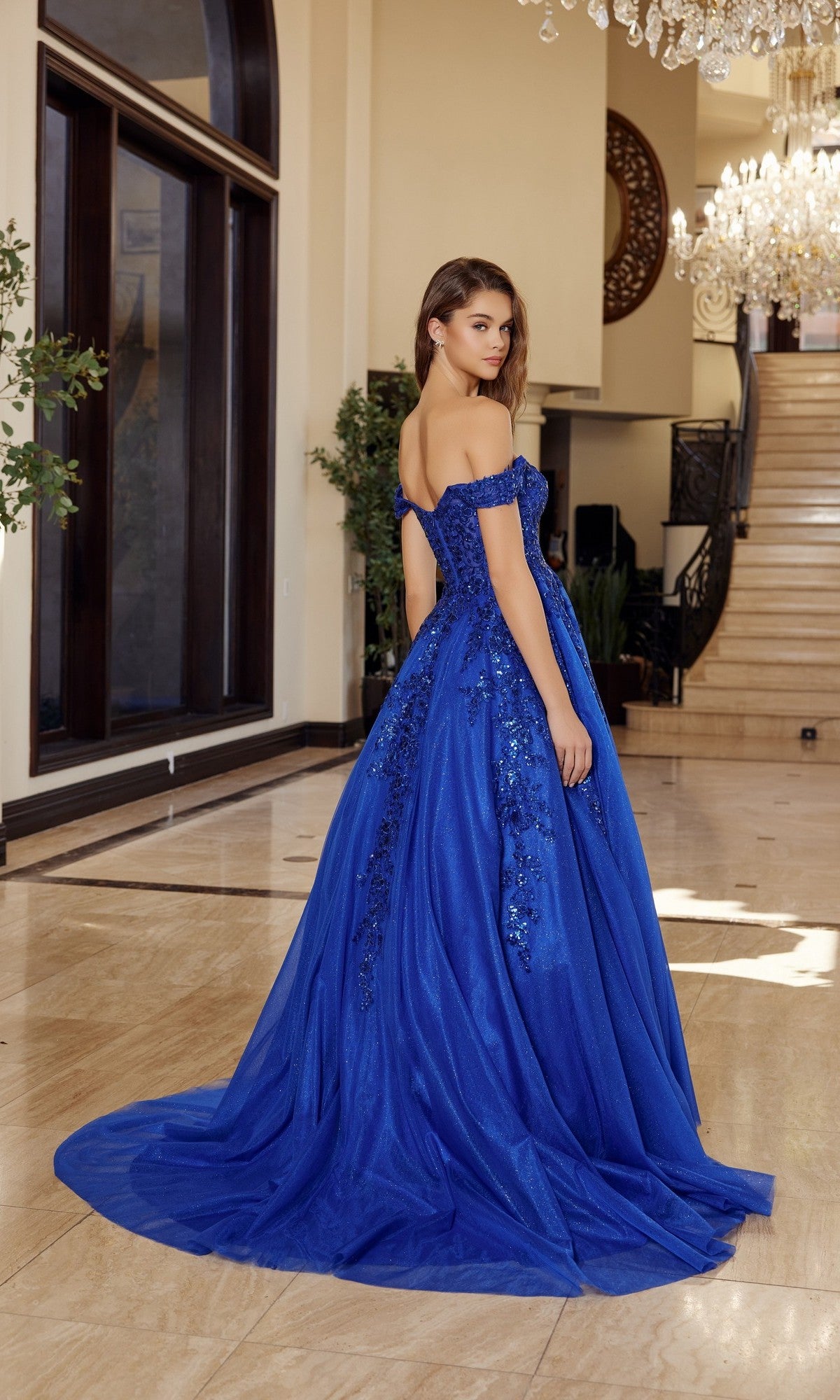 A woman stands indoors on a marble floor, facing away from a grand staircase, wearing the Nox Anabel H1881 Long Prom Dress—an off-the-shoulder tulle ball gown with a long train.